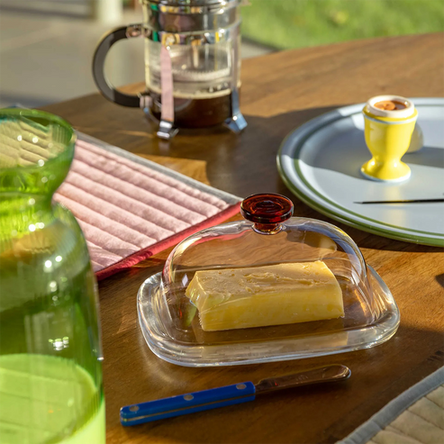Butter dish with a stick of butter on a wooden table with a French press and plates in the background.